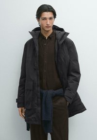 Sort dunjakke med hætte, forsynet med lynlås, store frontlommer og glat yderside. Modellen har en brun skjorte med knapper under.