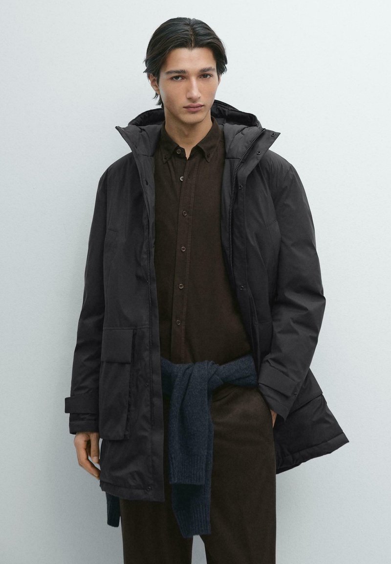 Sort dunjakke med hætte, forsynet med lynlås, store frontlommer og glat yderside. Modellen har en brun skjorte med knapper under.