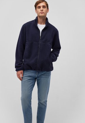 Marineblaue Fleecejacke mit Reißverschluss und Stehkragen, mit einer strukturierten Oberfläche und zwei Seitentaschen, kombiniert mit hellblauen Jeans.