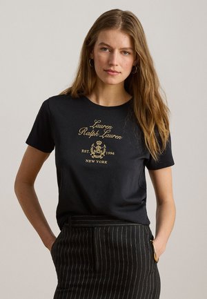 Pitkätukkainen nainen pukeutunut mustaan Lauren Ralph Lauren -t-paitaan, jossa on kultainen logo, ja mustaan neulospaitaan, seisoo kädet taskuissa.
