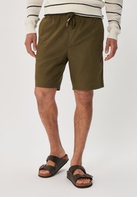 Olivové krátké kalhoty s pružným pasem a detailem na šňůrku. V kombinaci s hnědými sandály s dvojitými popruhy a korkovou podrážkou.