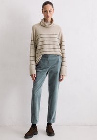 Beżowy sweter w paski z wysokim kołnierzem, zestawiony z zielonymi spodniami ze sztruksu i brązowymi butami wsuwanymi. Prosty, luźny fason z długimi rękawami.