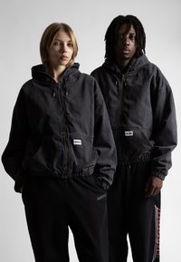 Juodi džinsiniai hoodie turi priekines užtrauktuką, elastines rankoves ir juosmenį. Asmenys dėvi suderintas juodas kelnes su subtiliu ženklinimu.