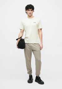 Férfi, bézs EA7 pólóban és melegítőnadrágban, fekete sportcipőben, fekete táskát tart egy egyszínű fehér háttér előtt.