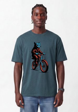Junger Mann mit geflochtenen Haaren, der ein blau-graues T-Shirt mit einer Grafik einer Katze trägt, die Fahrrad fährt.