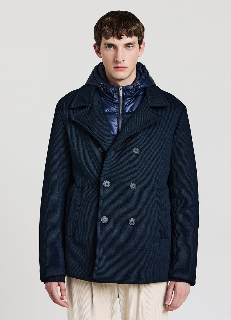 Marineblauer, zweireihiger Mantel mit strukturierter Stoffqualität, großen Revers und Vordertaschen, darüber getragen eine gesteppte, marineblaue Daunenjacke.