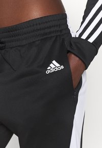 Personne portant un pantalon de survêtement noir Adidas avec des bandes blanches et un logo, main dans la poche zippée latérale, portant également une manche de veste assortie.