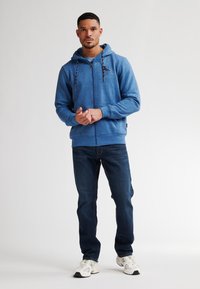 Felpa blu con cappuccio e zip, con lacci e logo, abbinata a jeans blu scuro e sneakers bianche. Texture liscia e vestibilità casual.