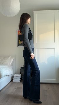 Dunkelblaue Schlagjeans, graues, geripptes Langarmshirt und eine kleine schwarze Handtasche. Der Raum verfügt über eine weiße Papierlaterne und einen Kleiderschrank.