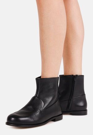 Paar schwarze Leder-Stiefeletten mit seitlichem Reißverschluss, getragen an nackten Beinen, stehend vor schlicht weißem Hintergrund.