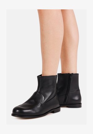Paire de bottines en cuir noir avec fermeture éclair latérale portées sur des jambes nues, posées sur un fond blanc uni.