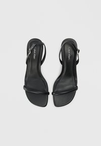 Black leather sandals with a minimal design, featuring two thin straps and an ankle buckle. Smooth texture and flat sole. Calvin Klein branding.
