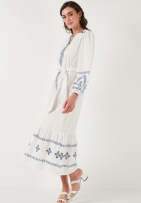 White long dress with blue embroidered accents on the sleeves and hem, featuring a V-neck, tie belt, and ruffled bottom. Smooth texture.