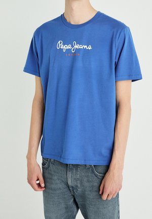 Mann trägt ein blaues Pepe Jeans London T-Shirt und ausgewaschene blaue Jeans, steht vor einem einfarbigen hellen Hintergrund.