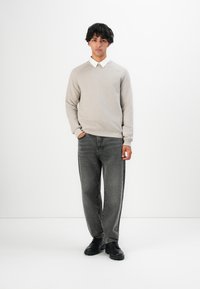 Jonge man die rechtop staat in een beige sweater over een witte blouse, vervaagde zwarte jeans en zwarte laarzen voor een effen witte achtergrond.