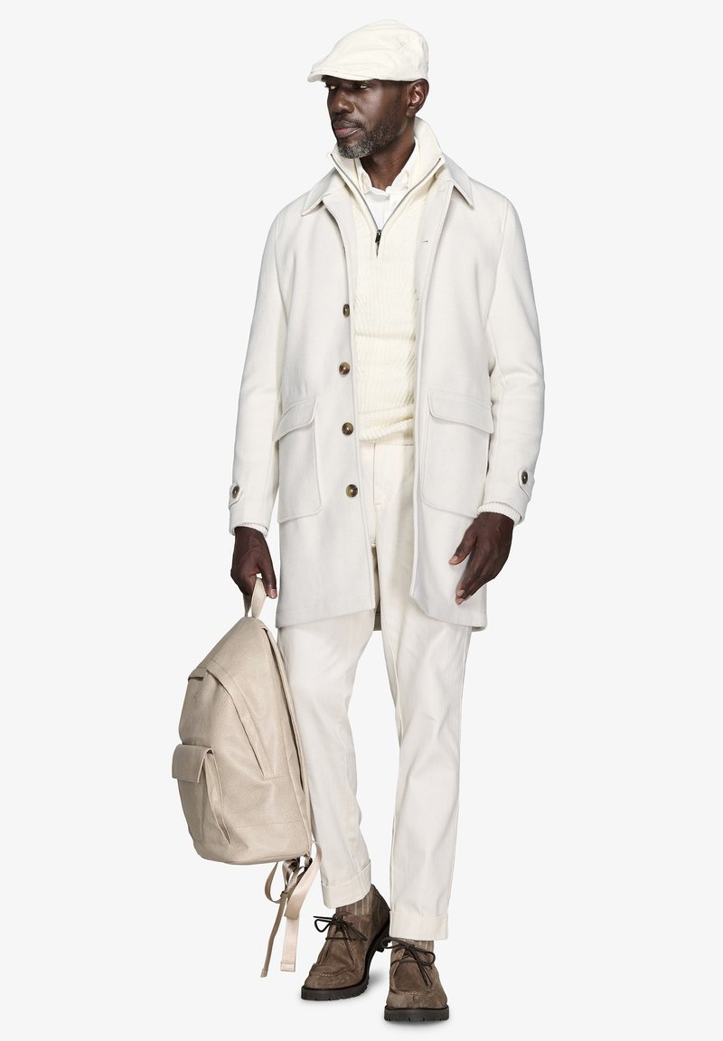 Abrigo de lana blanco con botones marrones, suéter de punto beige, mochila beige clara, pantalones claros y zapatos de cordones marrones. La gorra combina con el abrigo.