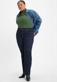 Jeans de ganga azul escuro, com uma cintura alta e um design de perna reta. Usados com um suéter de gola alta listrado em verde e uma jaqueta de ganga. Botas pretas de tornozelo.