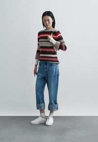 Gestrickter, gestreifter Pullover in Rot, Braun und Beige, kombiniert mit hochgekrempelten Jeans und weißen Slip-on-Schuhen mit goldenen Akzenten.