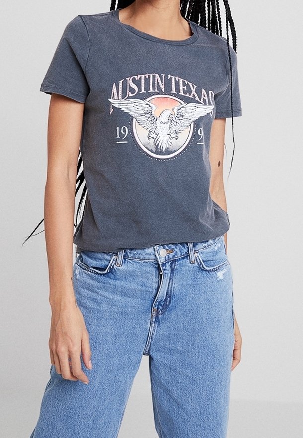 Camiseta de algodón gris con gráfico de "Austin Texas" y diseño de águila, cuello redondo, mangas cortas y corte relajado. Combinada con unos jeans de color azul claro.