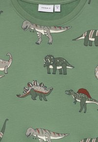 T-shirt verde in cotone con diversi dinosauri illustrati in tonalità di grigio e verde, con motivi sparsi e un colletto a costine.