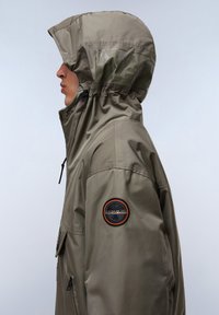 Oliegroene waterdichte jas met een hoge kraag, verstelbare capuchon en een ronde patch op de mouw. Heeft een gladde textuur en een rits.