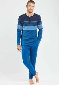 Homme debout pieds nus, portant un haut à rayures bleu à manches longues et un pantalon bleu uni, avec une main dans sa poche.