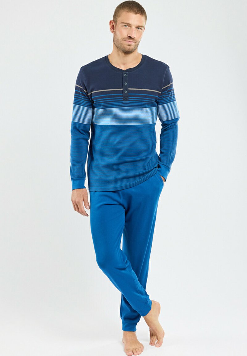 Homme debout pieds nus, portant un haut à rayures bleu à manches longues et un pantalon bleu uni, avec une main dans sa poche.