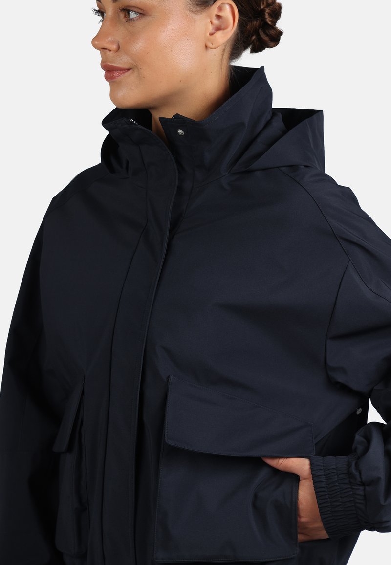 Ciemnoniebieska wodoodporna kurtka z wysokim kołnierzem, wieloma kieszeniami z przodu i elastycznymi mankietami dla pewnego dopasowania. Gładka faktura.