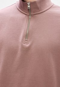 Světle růžový svetr s límcem na zip. Obsahuje měkkou texturu a logo "HUGO" vyražené na hrudi.