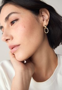 Gold-tone earrings featuring a twisted design intertwined with an elongated oval shape. The model has medium-length dark hair and a neutral top.