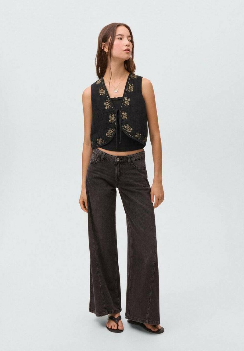 Črn skrajšan top z zlatimi vezeninami, v kombinaciji z visokim pasom temnimi kavbojkami. Model stoji v ravnih sandalih na svetli podlagi.