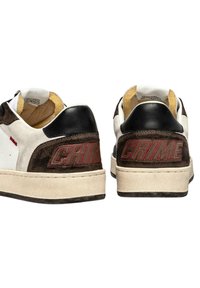 Un paio di sneakers bianche e marroni con toppe nere sul tallone, testo rosso "CRIME" sul retro e suole beige leggermente usurate, viste da dietro.