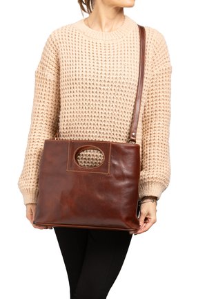 Persoon draagt een beige gebreide trui en houdt een grote bruine leren handtas met schouderriem en ovale uitsparing in het handvat vast.