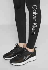 Zwarte sport sneakers met een textuurfinish, voorzien van witte accenten en veters, gecombineerd met zwarte leggings met het "Calvin Klein" logo.