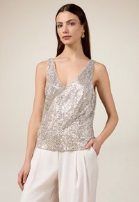 Top sin mangas de lentejuelas plateadas con un escote en V, diseño ajustado y textura suave, combinado con pantalones de pierna ancha en color crema.