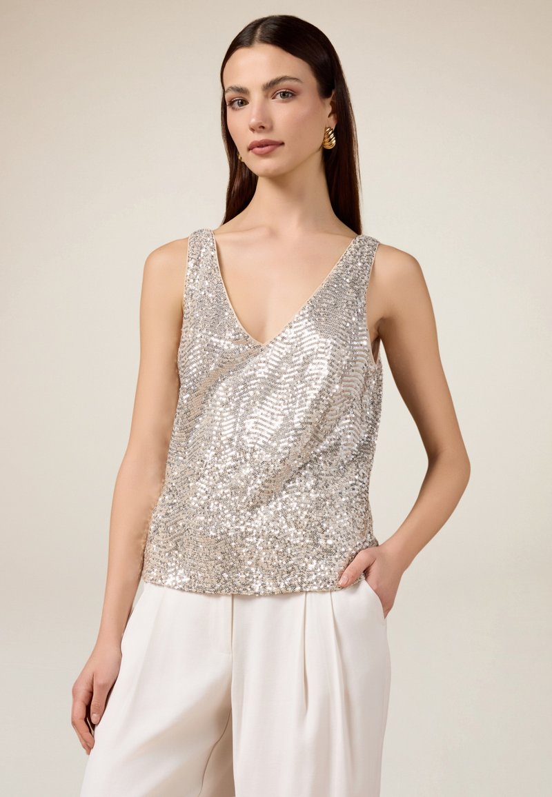 Top sin mangas de lentejuelas plateadas con un escote en V, diseño ajustado y textura suave, combinado con pantalones de pierna ancha en color crema.