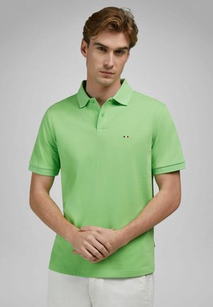 Jonge man met lichtbruin haar, gekleed in een felgroen poloshirt en witte broek, staand met gevouwen handen tegen een effen grijze achtergrond.