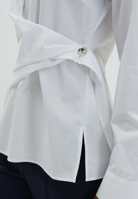 Weißes Baumwollhemd mit einem verdrehten Design an der Taille, gesichert durch einen silbernen Knopf. Verfügt über einen gebogenen Saum und seitliche Schlitze.