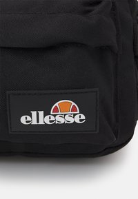 Czarna torba materiałowa o teksturowanej powierzchni, z prostokątną czarną metką z białym napisem i kolorowym logo.