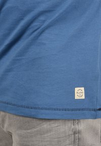 Modra bombažna majica s šivanim robom in blago oznako z napisom "DENIM BLEND with ATTITUDE" na spodnjem delu, v kombinaciji s sivo obleko.
