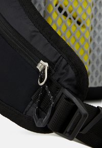 Pochette zippée en tissu noir avec des accents en mesh gris et un fond jaune. Inclut un crochet en métal, une sangle noire et un motif texturé.