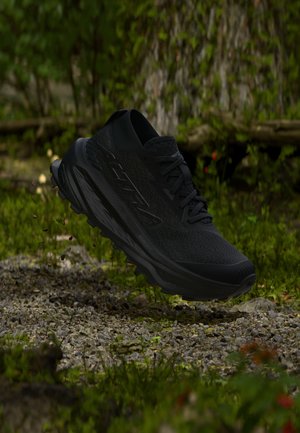 Zapatilla de correr negra en el aire sobre un camino de grava en un entorno de bosque verde con un tronco de árbol al fondo.