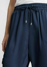 Pantalons légers bleu marine avec une taille élastique et un cordon de serrage. Caractéristiques en métal doré, poches côté et une texture lisse.