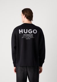 Černá mikina s dlouhými rukávy, na zadní straně s velkým textem a logem "HUGO". Hladký materiál, volný střih a rovný lem.
