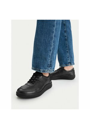 Stopy w czarnych skórzanych trampkach i niebieskich dżinsach stojące na jasnoszarej powierzchni.