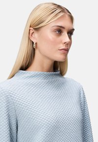Top blu chiaro con una texture in rilievo e colletto alto, caratterizzato da un motivo a rombi. La modella indossa piccoli orecchini a cerchio. Capelli lisci che incorniciano il viso.