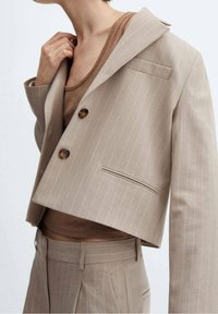 Blazer beige coupé rayé à chevrons avec un revers cranté, deux boutons et une poche poitrine unique, associé à un débardeur marron.