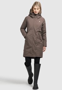 Rjav zimski dolg plašč s kapuco, ki ima žepke z zadrgo in je v kombinaciji z črnimi gumijastimi škornji do kolen. Gladka tekstura in eleganten dizajn.