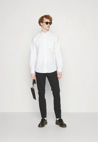 Hvit skjorte med knapper, svarte slanke bukser, svarte sko og solbriller. Skjorten har en brystlomme og en klassisk krage.