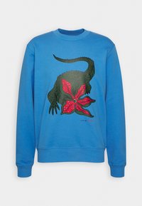 Sudadera azul con un gráfico de la cabeza de un reptil verde fusionada con una estrella de mar roja, con los logotipos de Lacoste y Netflix debajo del diseño.